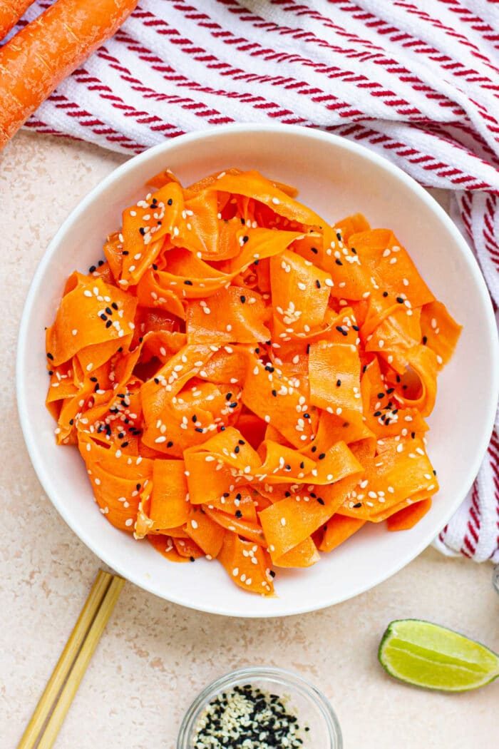 A refreshing bowl of raw carrot salad features delicate ribbons topped with sesame seeds, resting beside a striped towel and a zesty lime wedge.