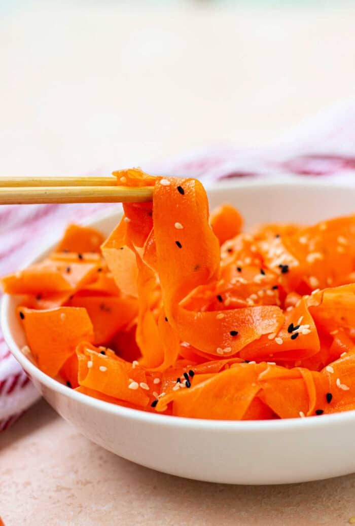 A bowl of raw carrot salad featuring delicate ribbons sprinkled with sesame seeds, gracefully lifted with chopsticks.