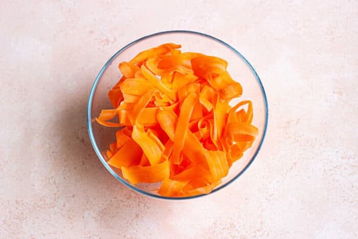 A glass bowl brimming with raw carrot salad featuring vibrant orange ribbons, set against a light beige background.