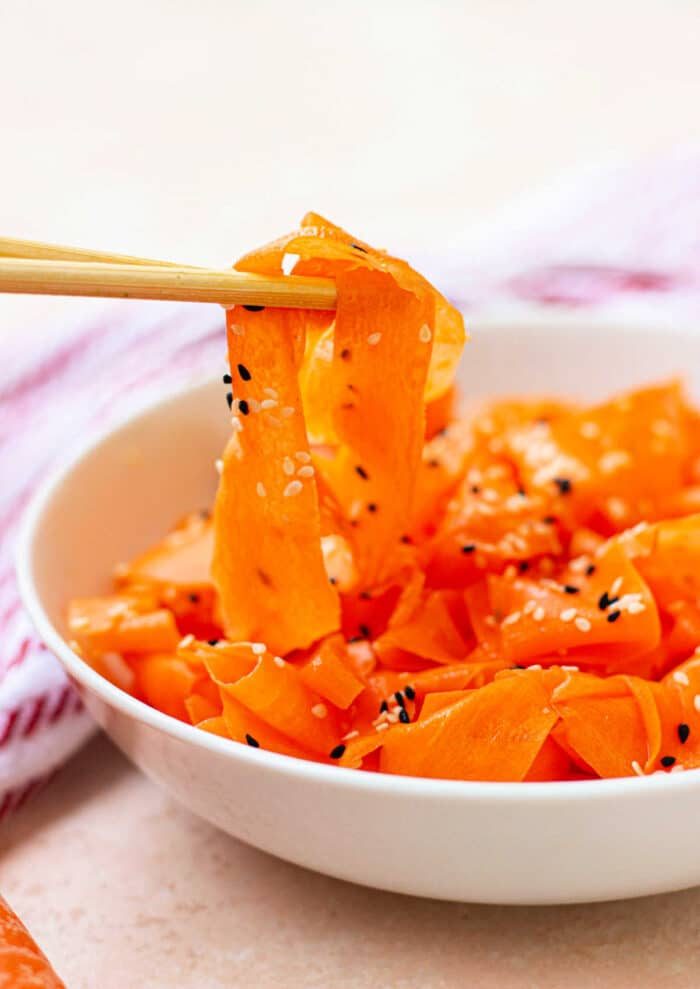 Chopsticks gracefully hold pieces of a good Raw Carrot Salad, sprinkled with sesame seeds, freshly lifted from a pristine white bowl.