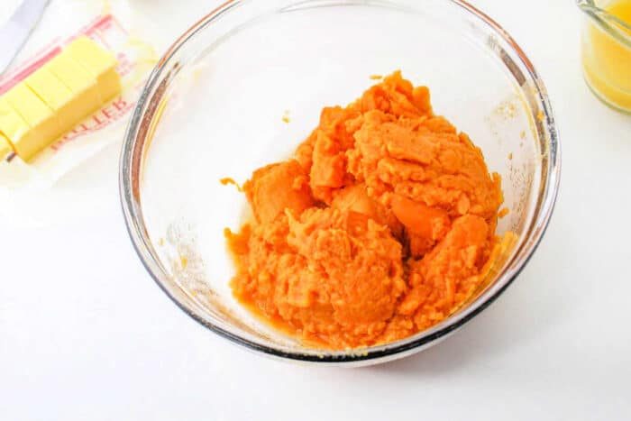 A glass bowl filled with mashed sweet potatoes is on a white surface. Next to it, there is a package of partially sliced butter and a small jar containing a yellow liquid, solid ingredients for your recipe for Sweet Potato Pie with Sweetened Condensed Milk.