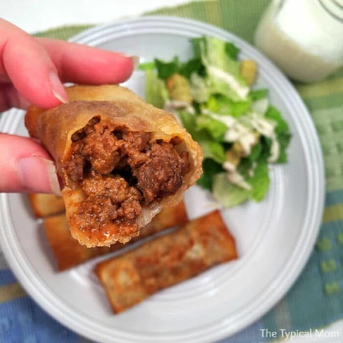 Hand holding a crispy egg roll with ground beef filling over a plate of egg rolls and salad—solid inspiration for recipes using egg roll wrappers.