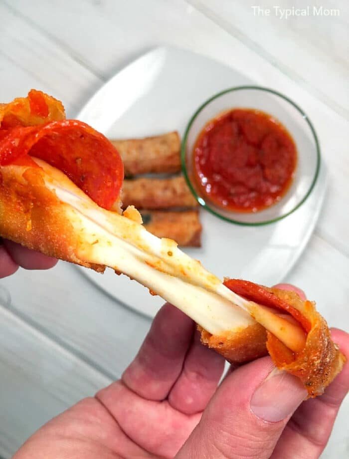 A hand holds a cheesy pepperoni pizza stick made with egg roll wrappers, with sauce and more sticks on a plate in the background—one of the tastiest recipes using egg roll wrappers.