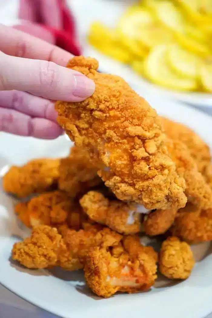 A hand holds a crispy air fried chicken tender over a plate piled with more golden tenders, fresh as if straight from the fryer—ideal for reheating chicken tenders in an air fryer.