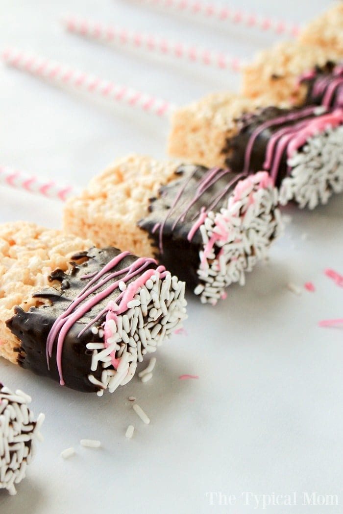 Close-up of rice krispy treat pops on sticks, each partially dipped in chocolate and adorned with pink and white drizzles and sprinkles. These delightful treats are neatly arranged in a line on a pristine white surface.