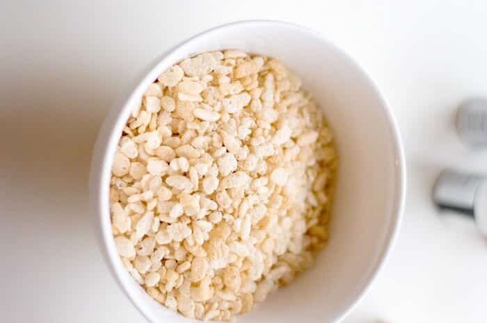 A white bowl filled with puffed rice cereal is placed on a white surface. The cereal is light, airy, and crisp, reminiscent of the texture in rice krispy treats. The edge of another object is slightly visible in the blurred background.
