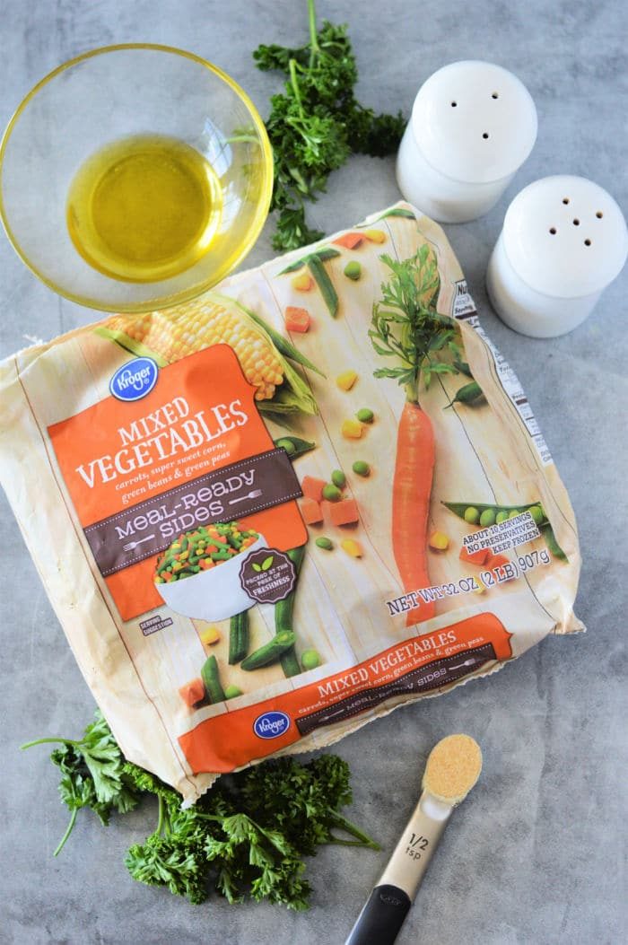 A package of mixed vegetables, ideal for roasting frozen vegetables, features a carrot and corn design on a gray surface. Nearby are a bowl of oil, parsley sprigs, a half teaspoon of garlic powder, a spoon, and salt and pepper shakers.