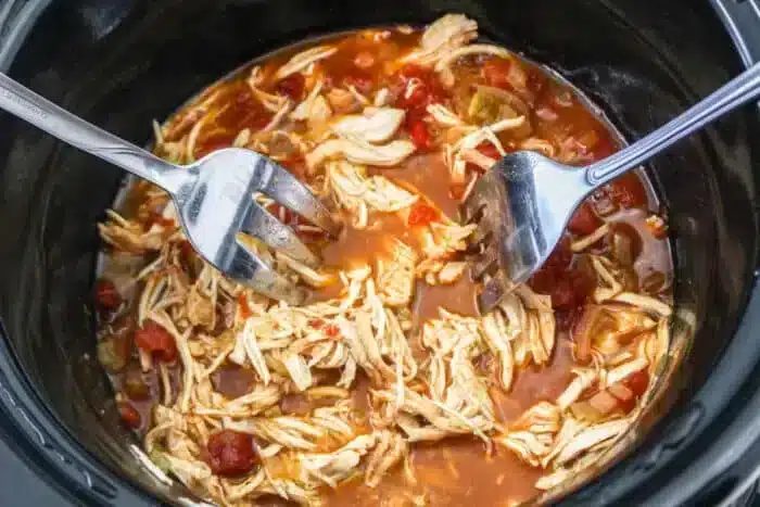 Two forks are shredding cooked chicken in a slow cooker. The chicken mingles with a vibrant Rotel sauce featuring diced tomatoes and green peppers, solid for mouth-watering tacos.