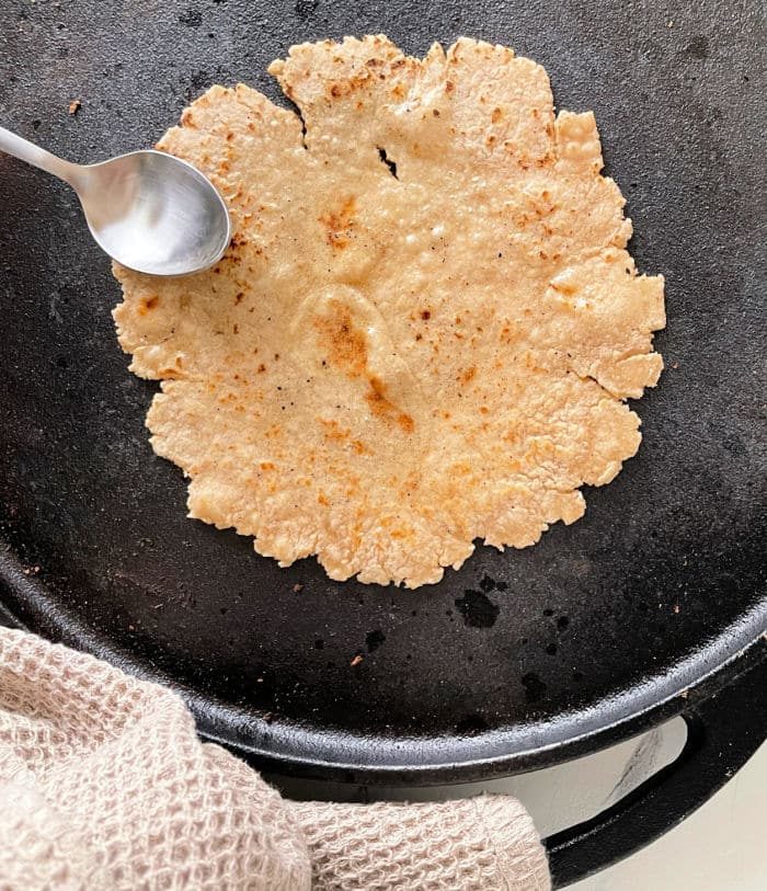 Roti cooking on a skillet with a spoon, next to a textured cloth, offers a snapshot of how to make roti perfectly at home.