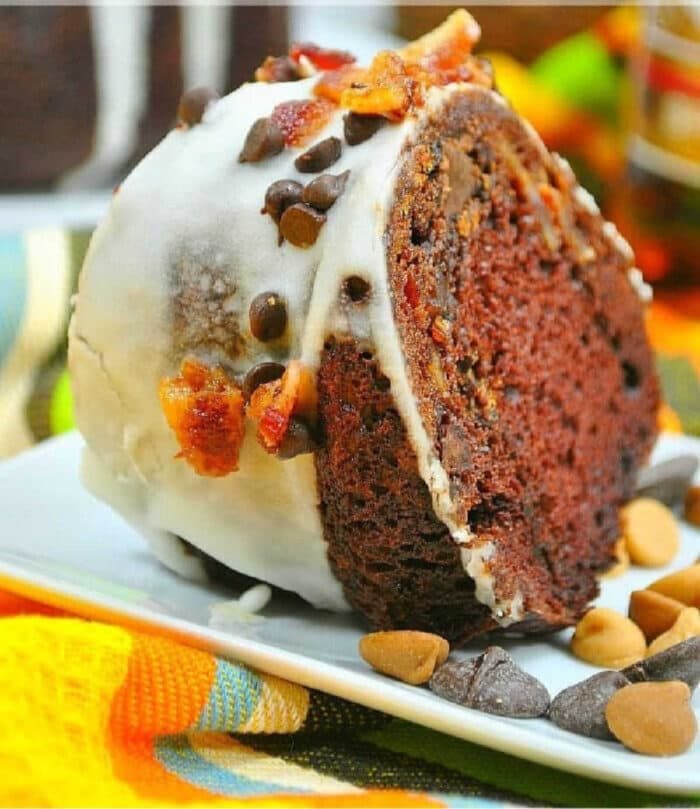 Slice of cake mix chocolate rum bundt cake with white icing, chocolate chips, and bacon bits on a white plate.