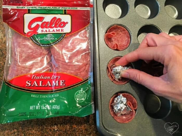 A hand presses foil into salami slices in a muffin tin to form savory salami cups; a pack of Gallo salame sits beside the tray.