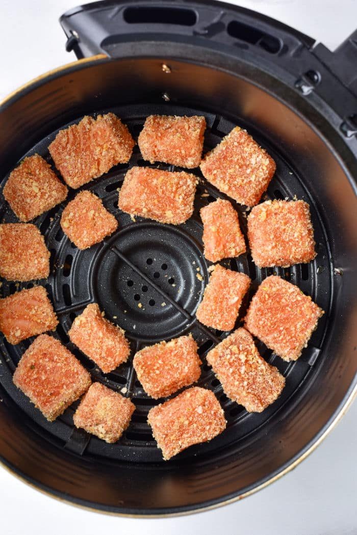 Breaded salmon pieces, similar to delectable salmon bites, sit in an air fryer basket, ready to be cooked.