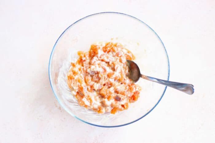 A transparent bowl containing a mixture of small, salmon chunks in a creamy white dressing is placed on a light surface, reminiscent of deconstructed Salmon Sushi Cups. A metal spoon rests inside the bowl.