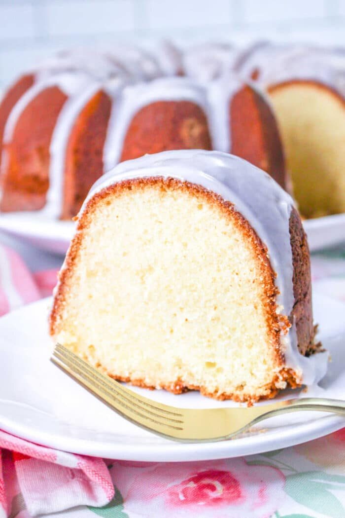 A slice of million dollar Pound Cake glazed bundt cake sits on a white plate with a gold fork, with the rest of the good cake in the background.