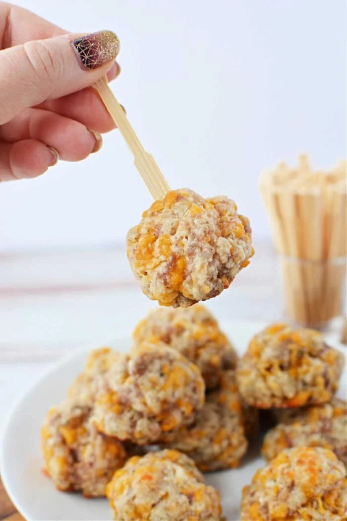 A hand holds a wooden pick with a cheese and sausage ball above a plate of delectable creations made using Red Lobster Biscuit Mix, enhancing the savory flavors of classic sausage balls.