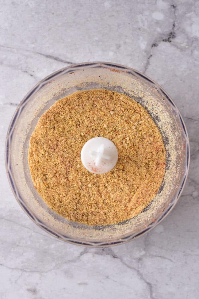 Food processor bowl with finely ground seasoned breadcrumbs on a light gray marble countertop.