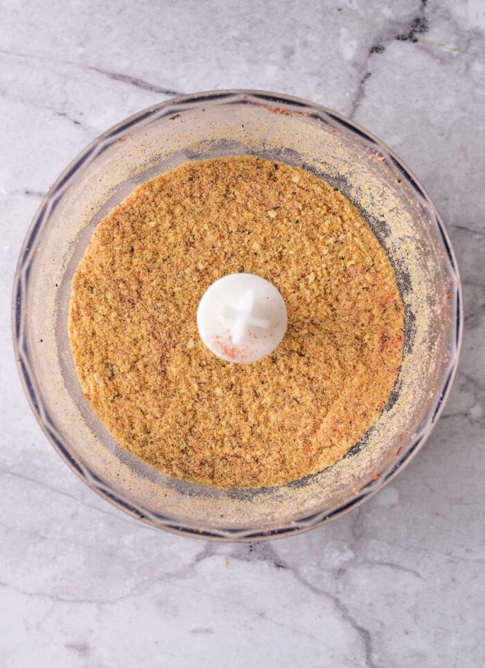 Seasoned breadcrumbs in a food processor bowl on a light marble countertop, viewed from above.