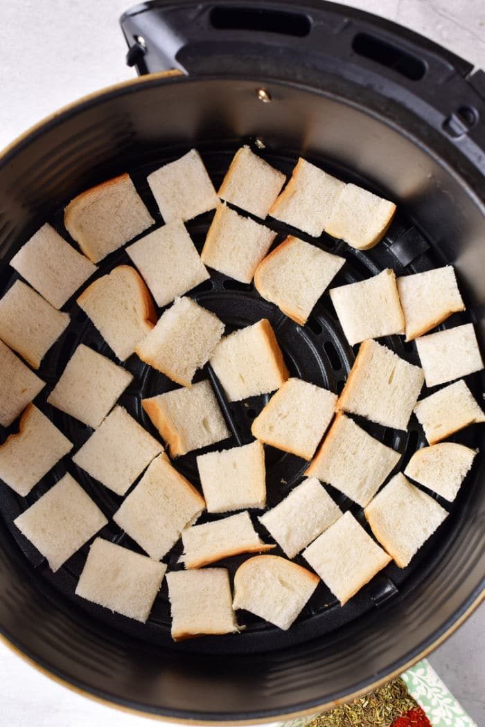 Cubed bread pieces spread evenly in an air fryer basket, solid for making seasoned breadcrumbs or toasting until golden brown.