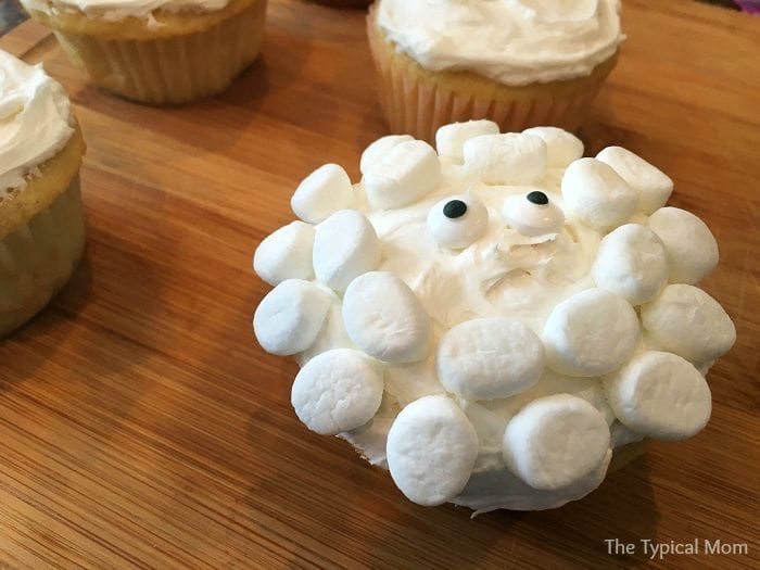 Sheep cupcakes, adorned with white frosting and mini marshmallows to perfectly mimic a sheep's face, rest charmingly on a wooden surface.