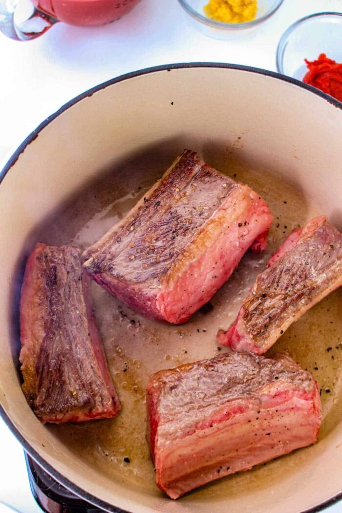 Four pieces of browned short rib meat are cooking in a large pot, solid for tacos. The meat has a seared exterior. In the background, there are small bowls with ingredients, including a bright red and yellow substance. The pot and ingredients are set on a white surface.