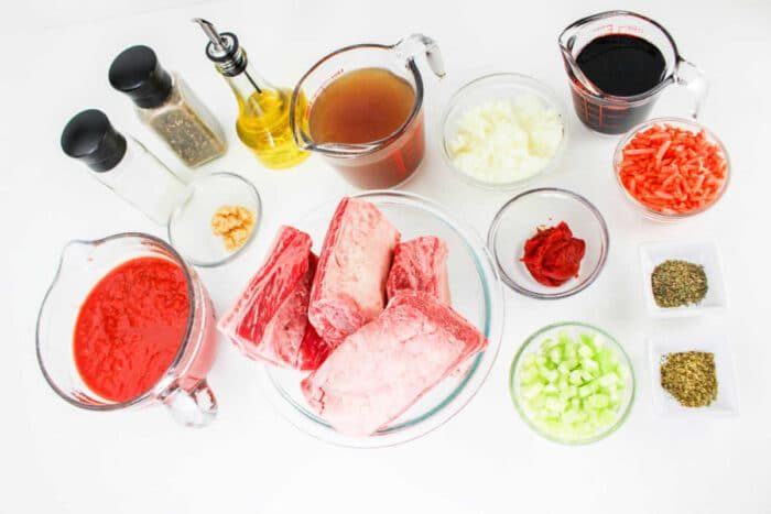 A collection of ingredients for a recipe arranged on a white surface, including raw beef short ribs solid for Short Rib Tacos, tomato sauce, beef broth, diced onions, shredded carrots, chopped celery, red wine, tomato paste, minced garlic, cooking oil, salt, pepper and dried herbs.