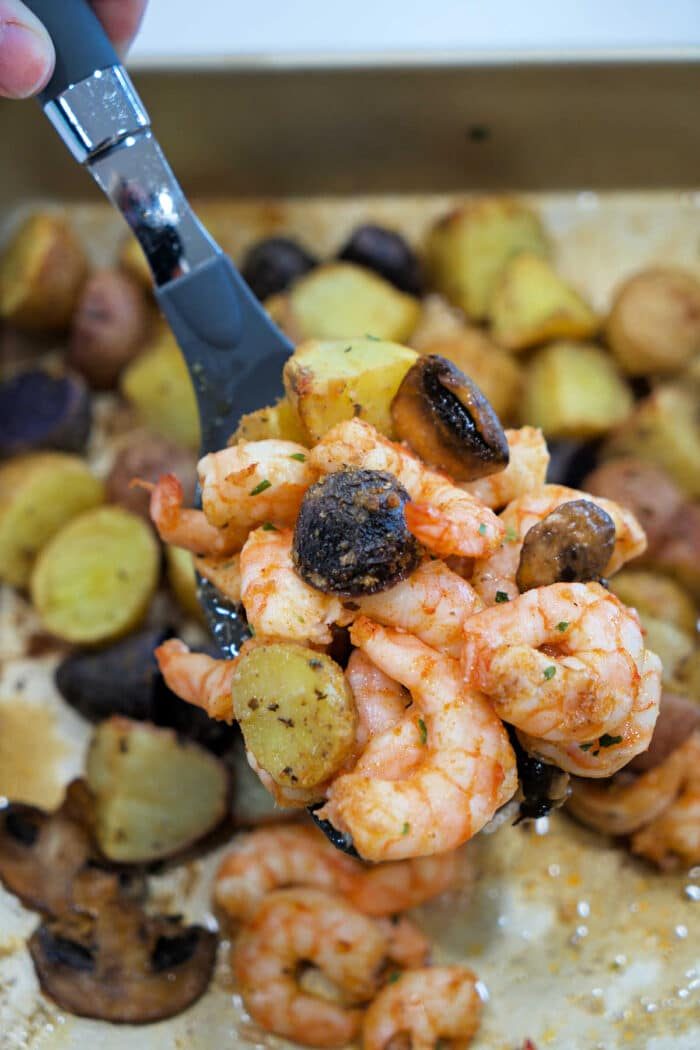 A spoon holds shrimp and potatoes with mushrooms above a tray filled with roasted shrimp and potatoes, plus savory mushrooms.