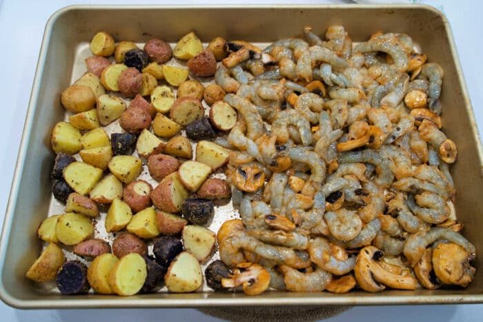 Sheet pan with seasoned baby potatoes on the left and raw shrimp and mushrooms on the right, this shrimp and potatoes combo is ready to bake.