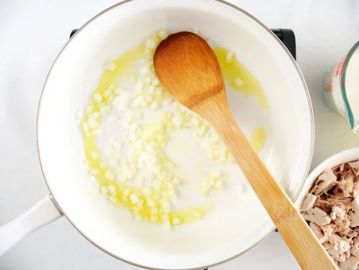 Diced onions and oil sizzle in a white pan with a wooden spoon, next to a bowl of shredded meat—solid for starting your simple chicken pot pie recipe.