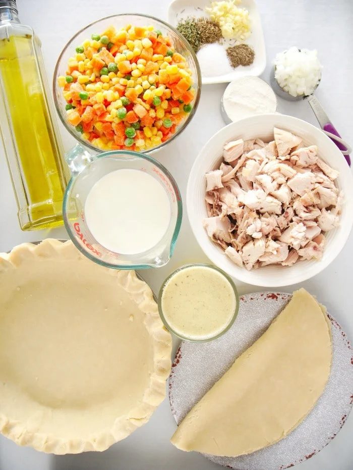 Ingredients for a simple chicken pot pie recipe, including vegetables, chicken, pie crust, milk, spices, and olive oil, laid out on a table.