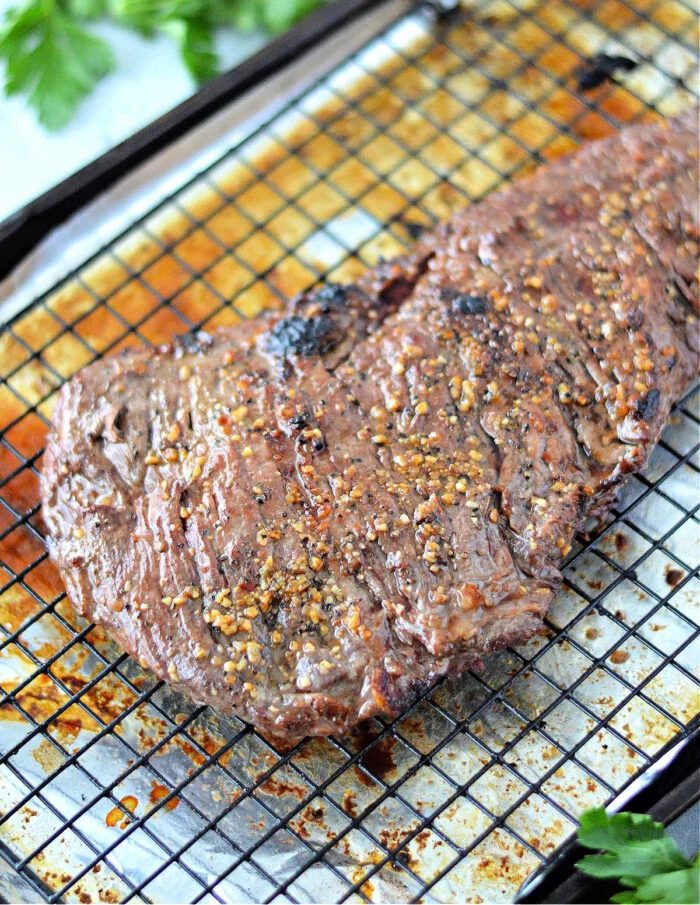 A cooked, seasoned skirt steak in oven rests on a wire rack over a baking sheet, with parsley leaves nearby.