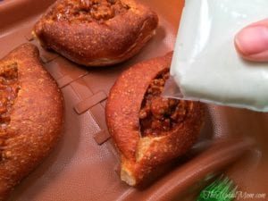 Close-up of a hand pouring white sauce from a bag onto three baked sloppy joe bread bowls filled with cooked minced meat on a brown tray.