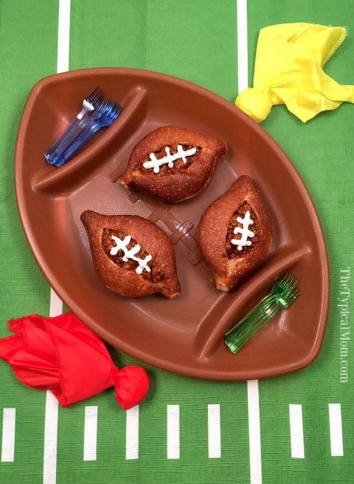 A brown football-shaped platter holds three sloppy joe filled bread bowls with sour cream lacing on the top.