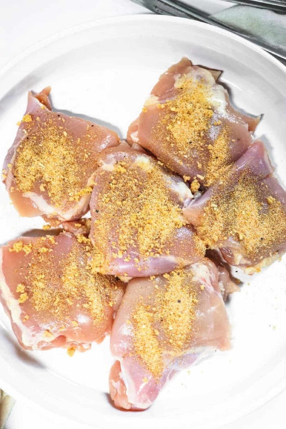 Raw chicken thighs on a white plate, sprinkled with a yellow seasoning powder—solid for making tender Slow Cooker BBQ Chicken Thighs.