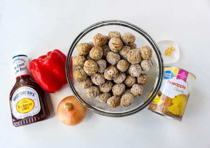 A bowl of frozen meatballs, solid for Slow Cooker BBQ Meatballs, is surrounded by a bottle of barbecue sauce, a red bell pepper, an onion, a can of pineapple chunks, and minced garlic on a white surface.