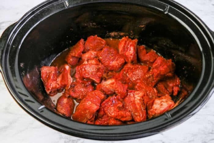 Chunks of raw beef coated in marinade inside a black slow cooker, ready to become tender slow cooker beef barbacoa on a marble surface.