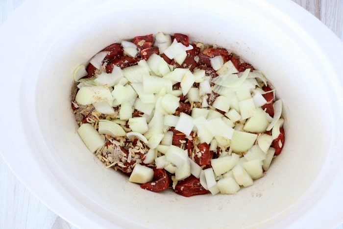 Chopped onions and spices mingle with slow cooker beef tips in a white slow cooker.