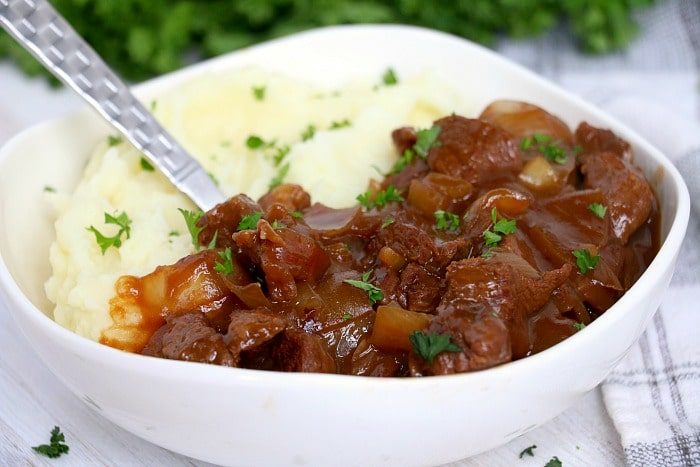 A bowl of creamy mashed potatoes and slow cooker beef tips stew garnished with chopped parsley, with a fork resting inside.