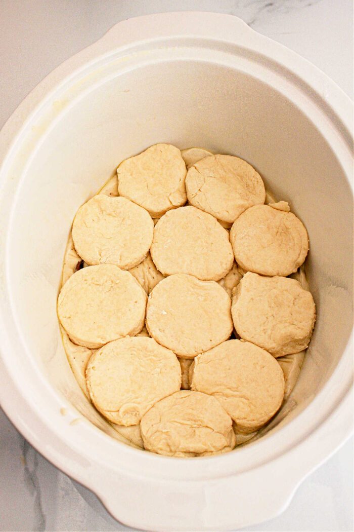 A white slow cooker with a lid open, showcasing an arrangement of what appears to be raw dough biscuits neatly placed inside. The interior and the sides of the slow cooker are visible. The background shows a grayish-white surface, setting the stage for some good Slow Cooker Biscuits and Gravy.