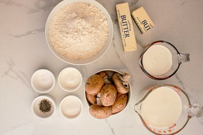 A top-down view of cooking ingredients on a white marble surface. There are flour, two sticks of butter, milk, cream, whole mushrooms, and small bowls containing salt, pepper, and sugar—solid essentials for preparing Slow Cooker Biscuits and Gravy.
