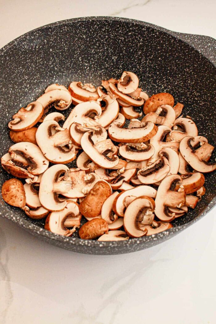 A black, speckled frying pan filled with slices of raw mushrooms sits invitingly on a light-colored countertop, setting the solid stage for a hearty Slow Cooker Biscuits and Gravy breakfast.