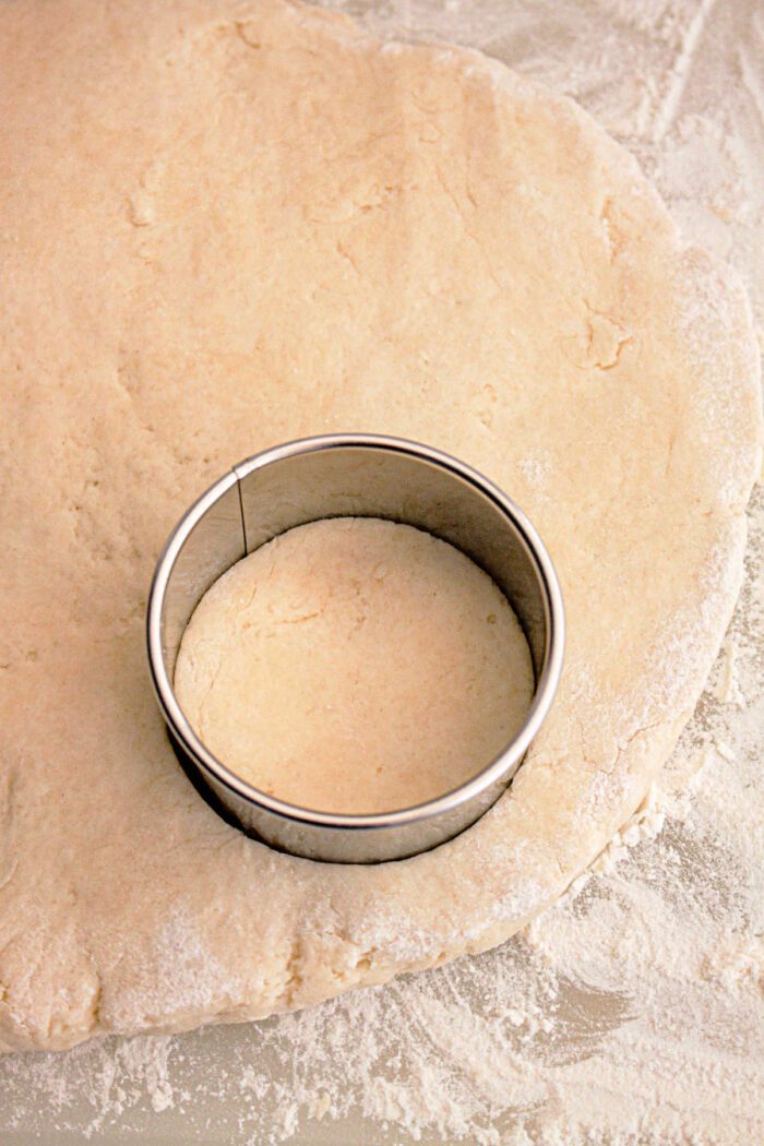 A metal round cookie cutter is pressed into rolled-out dough on a floured surface, creating a solid shape for Slow Cooker Biscuits and Gravy.