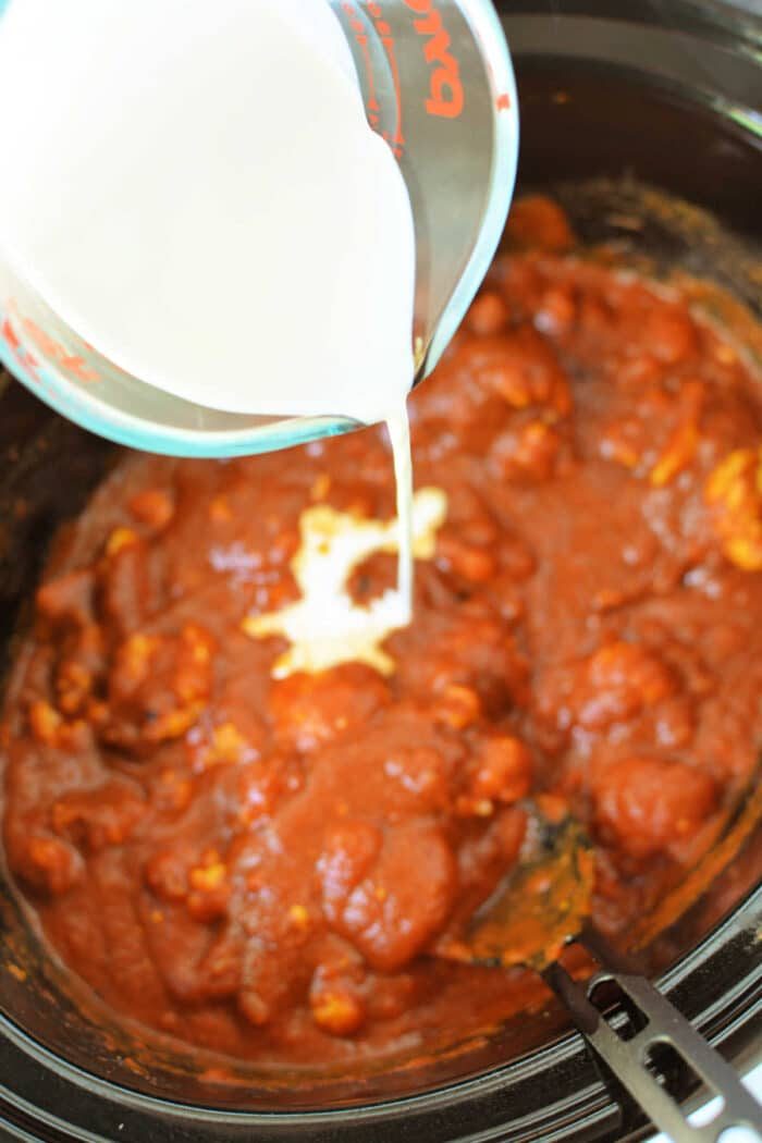 Milk is being poured from a measuring cup into a slow cooker filled with a tomato-based sauce, solid for making Slow Cooker Chicken Makhani.