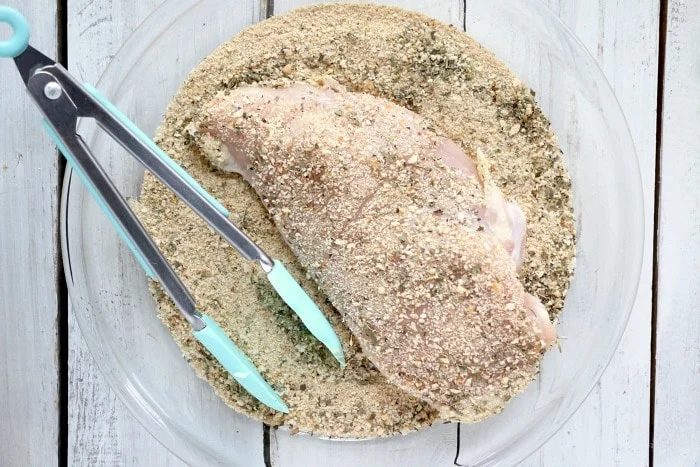 Raw chicken breast being coated in seasoned breadcrumbs with tongs in a glass dish on a white wooden surface, preparing for a good slow cooker chicken parmesan.