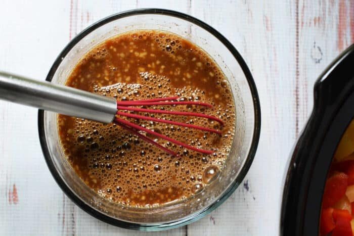 A metal whisk with a red handle mixes a brown sauce—solid for slow cooker chicken—in a glass bowl on a white wooden surface.