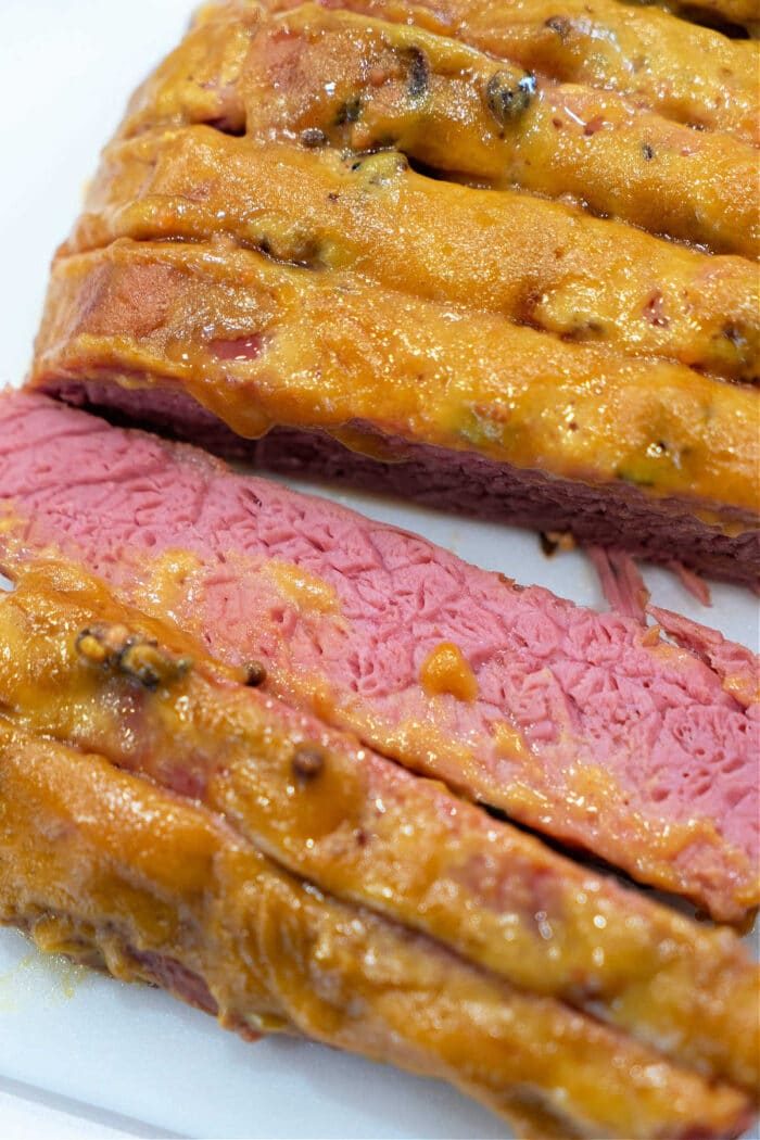 Sliced slow cooker corned beef brisket topped with a melted golden cheese and mustard mixture on a white surface.