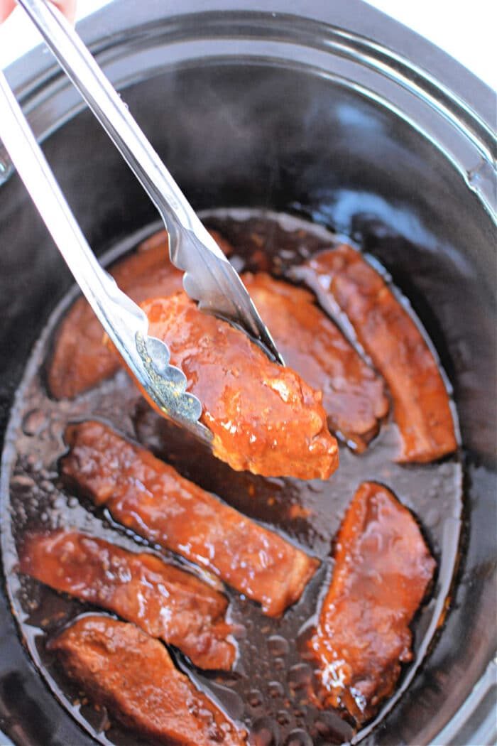 Tongs holding saucy, cooked country ribs hover over a slow cooker brimming with country-style ribs.