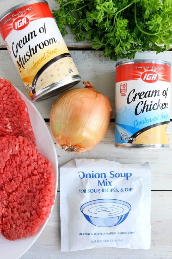 Slow cooker cube steak ingredients: ground beef, an onion, parsley, two cans of soup, and onion soup mix on a white wooden surface.
