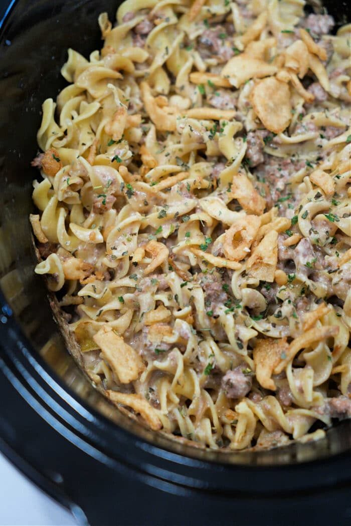 Close-up of creamy Ground Beef Stroganoff noodle casserole with herbs and crispy onions in a black slow cooker.