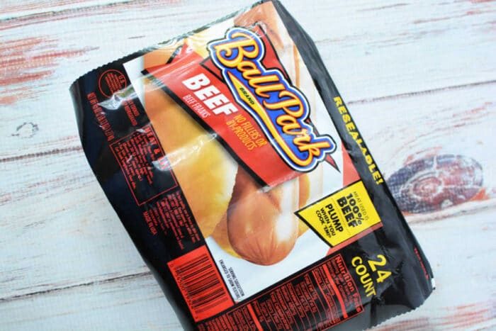 A package of Ball Park brand beef hot dog buns sits on a wooden surface. The packaging shows hot dogs and reads "BEEF - Made with 100% Beef Hot Dogs." solid for making slow cooker hot dogs, the package contains 24 individually wrapped hot dogs.