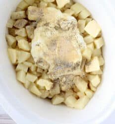 Diced potatoes with butter, cream, and seasonings in a white slow cooker, ready to be cooked into creamy slow cooker mashed potatoes.